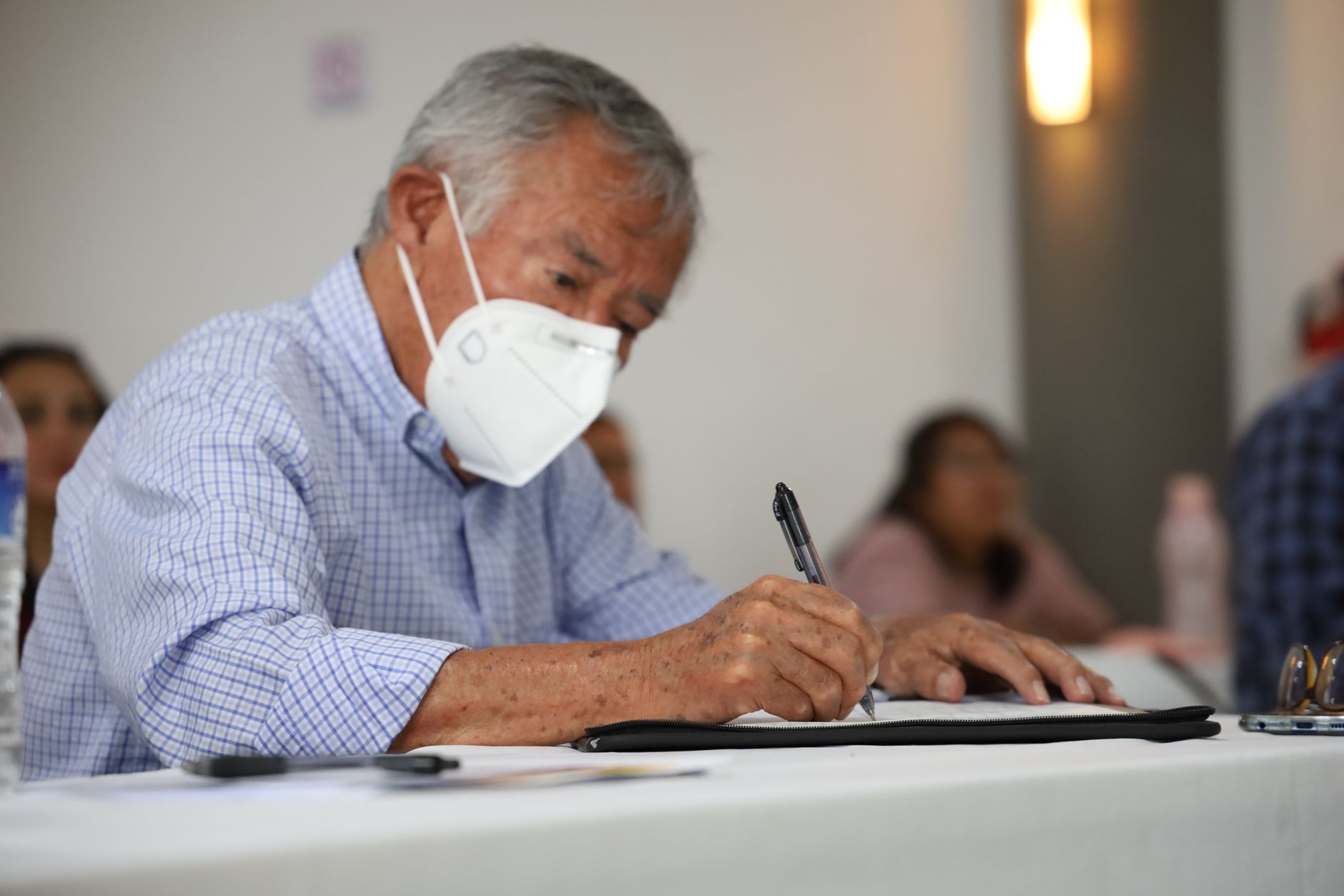 Elderly man with face mask sitting indoors, focused on writing in a notebook.