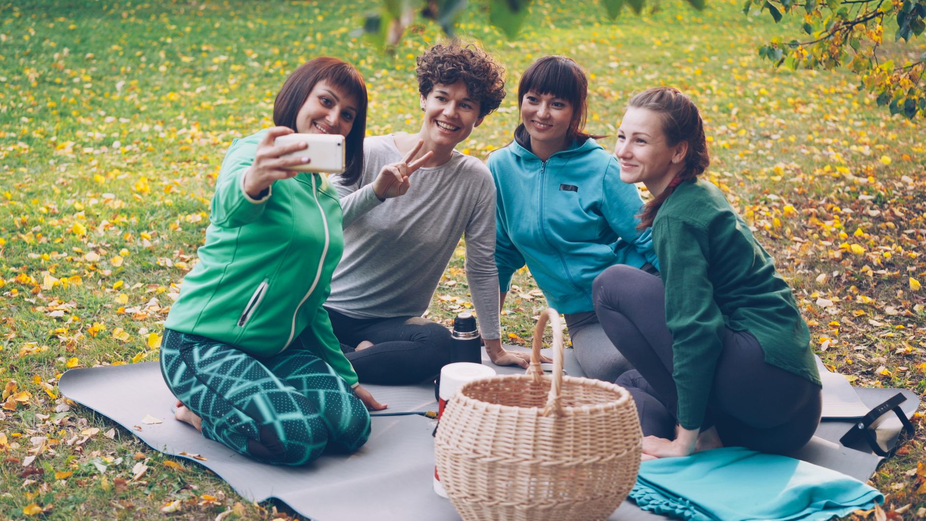 Four women taking a selfie during a yoga and picnic session in a park with autumn leaves.