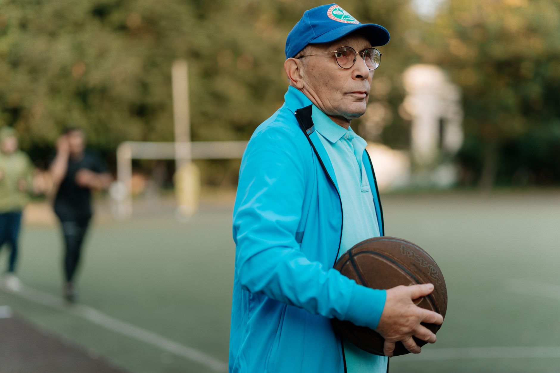 Elderly man with glasses and cap holding a basketball on an outdoor court.