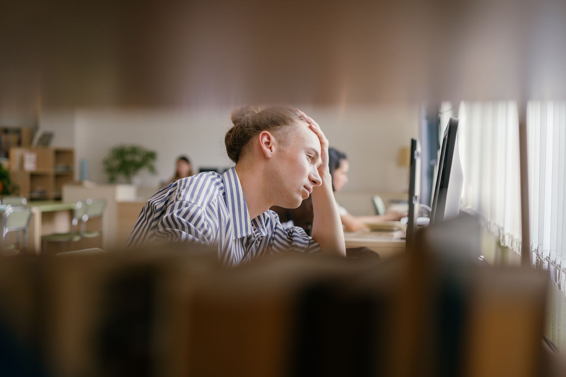 A student appears stressed while studying in a library, focusing on a computer screen.
