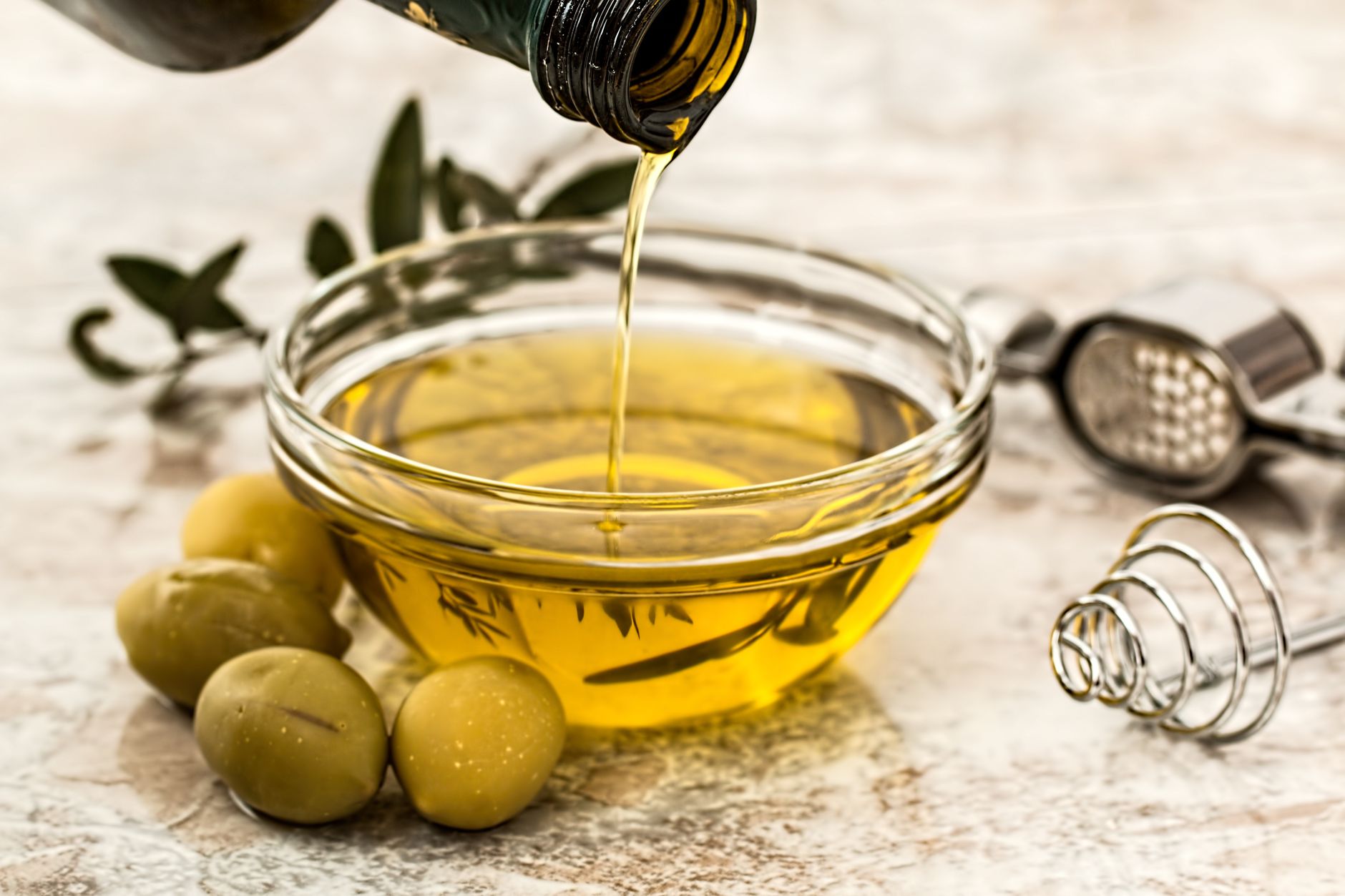 Close-up of olive oil being poured into a glass bowl surrounded by fresh olives and kitchen tools.
