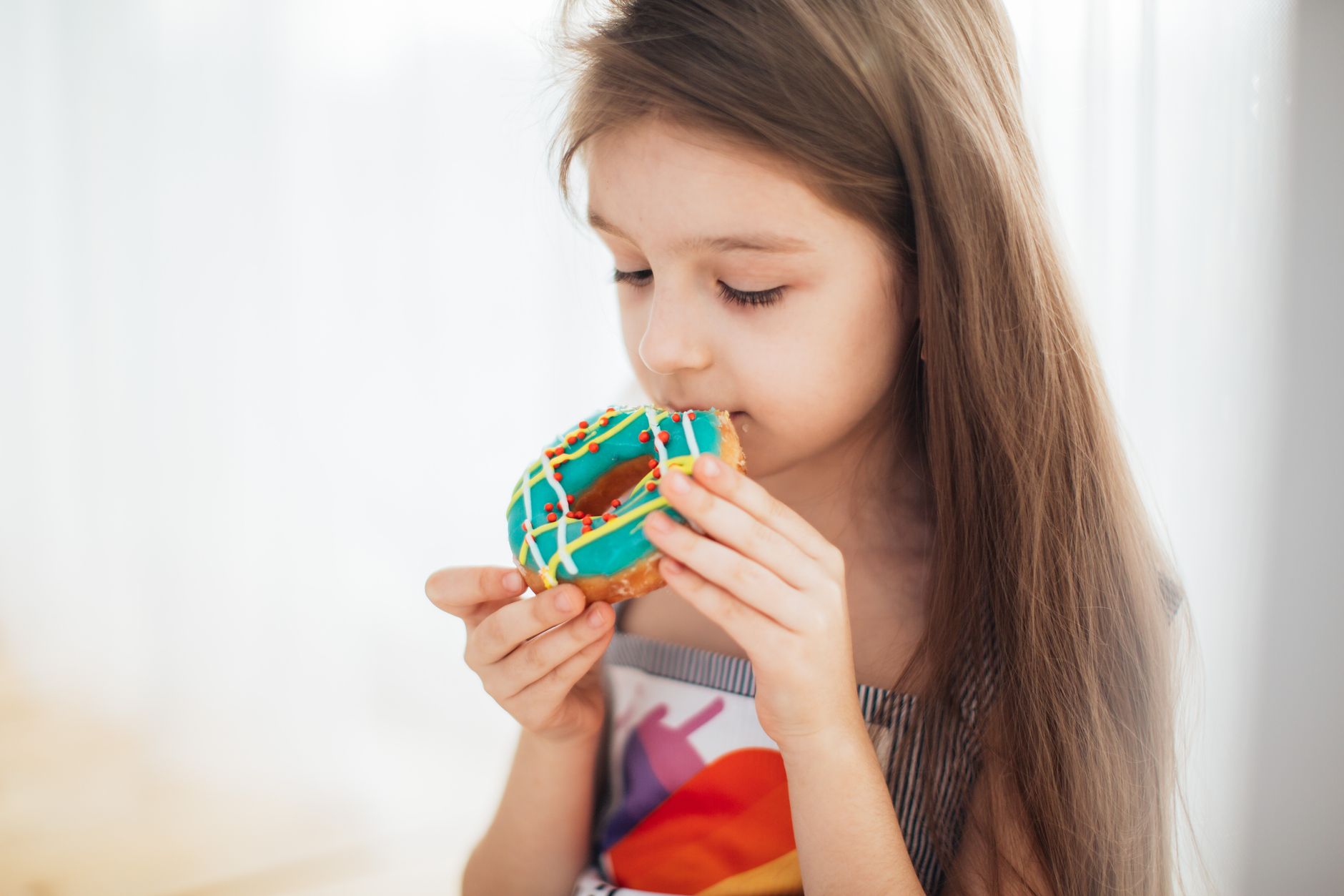 A young girl savors a vibrant donut, indoors during the daytime, depicting joy and sweetness.