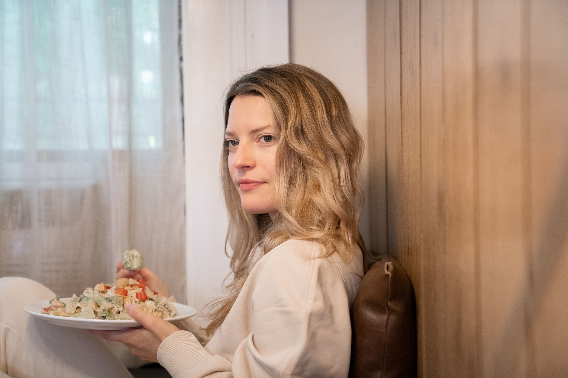 A woman with blonde hair enjoys a healthy salad while sitting at home indoors, exuding comfort and relaxation.