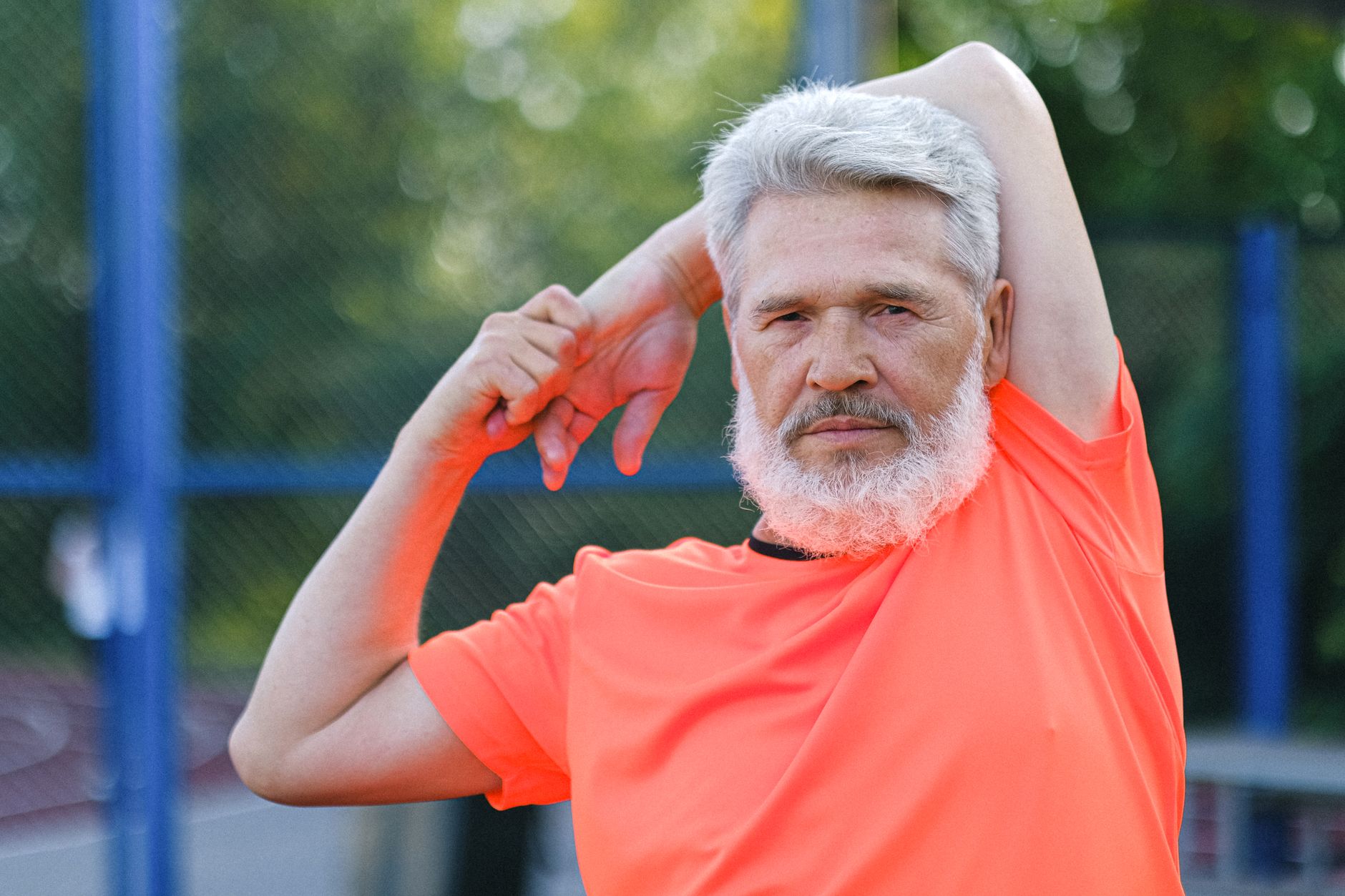 Confident bearded male with gray hair in sportswear stretching arms during workout on sports ground and looking at camera