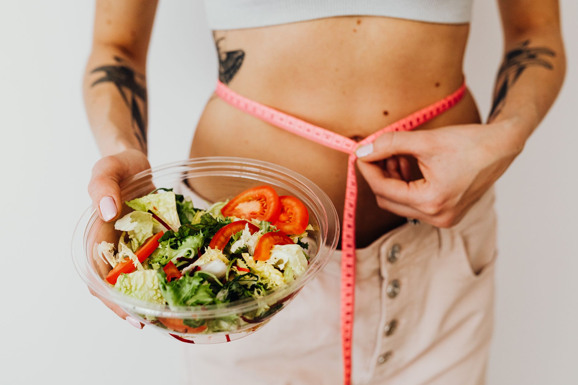 Woman holding vegetable salad and measuring waist with tape, symbolizing healthy lifestyle choices.