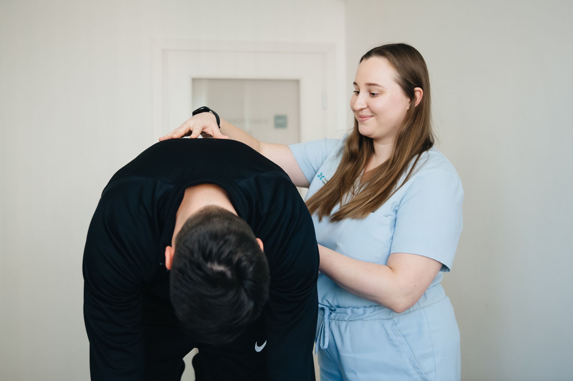 A physiotherapist smiling while assisting a patient with a back exercise in a clinic.