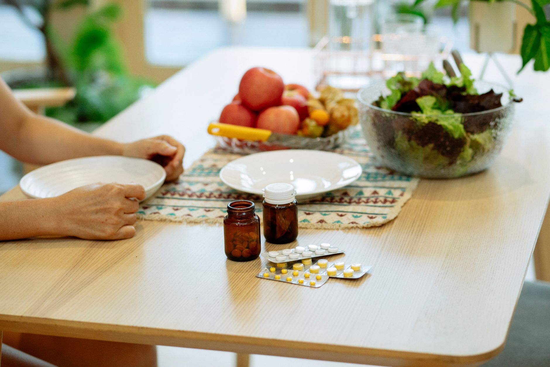 From above of crop anonymous person sitting at table with pile of various medicines and bowls of fresh lettuce salad and ripe fruits in kitc