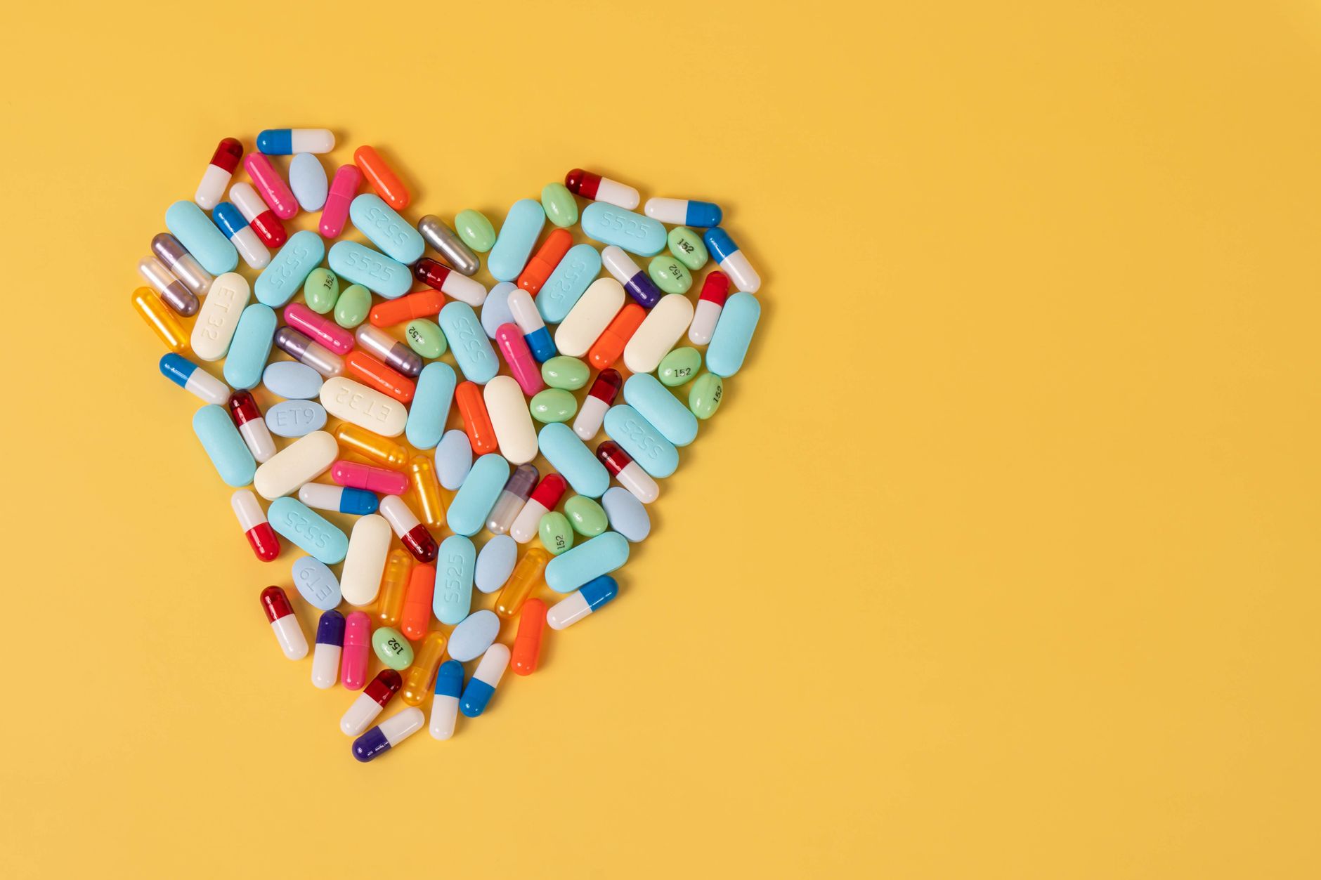 A heart-shaped cluster of multicolored pills on a vibrant yellow background, symbolizing healthcare and medicine.