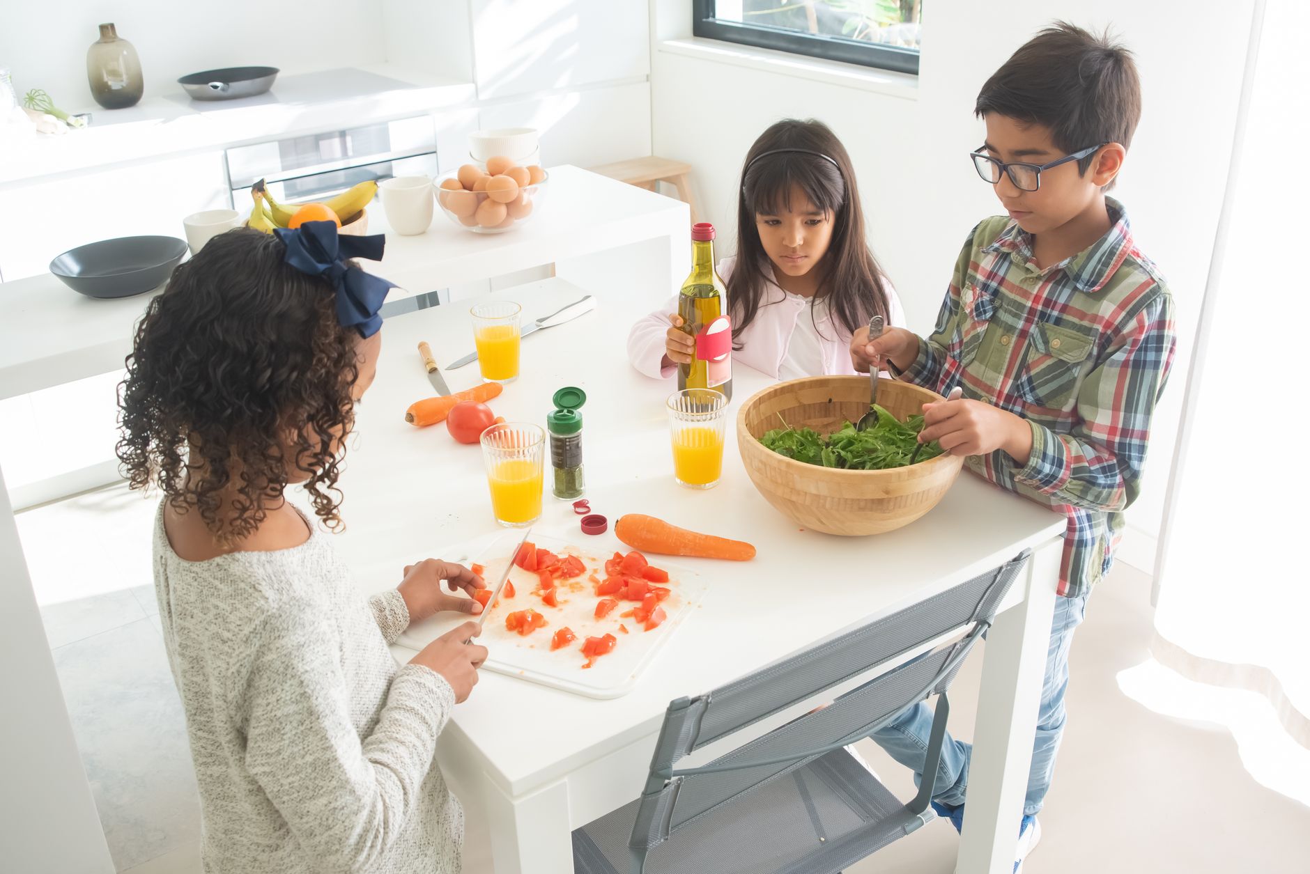 Three children preparing a fresh vegetable salad together in a bright kitchen.