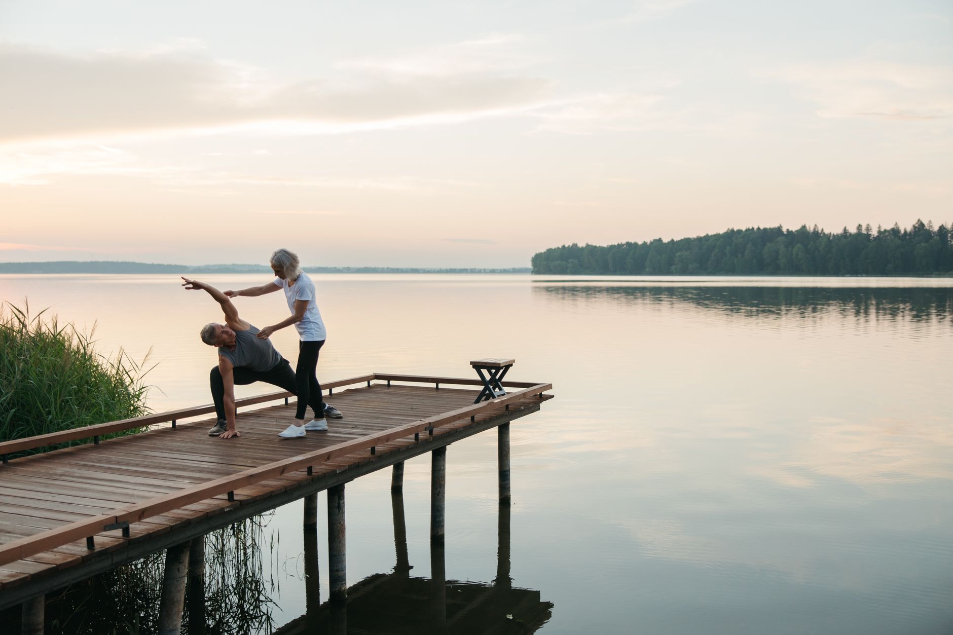 Senior adults practicing yoga on a wooden dock by a serene lake during sunrise, promoting health and mindfulness.