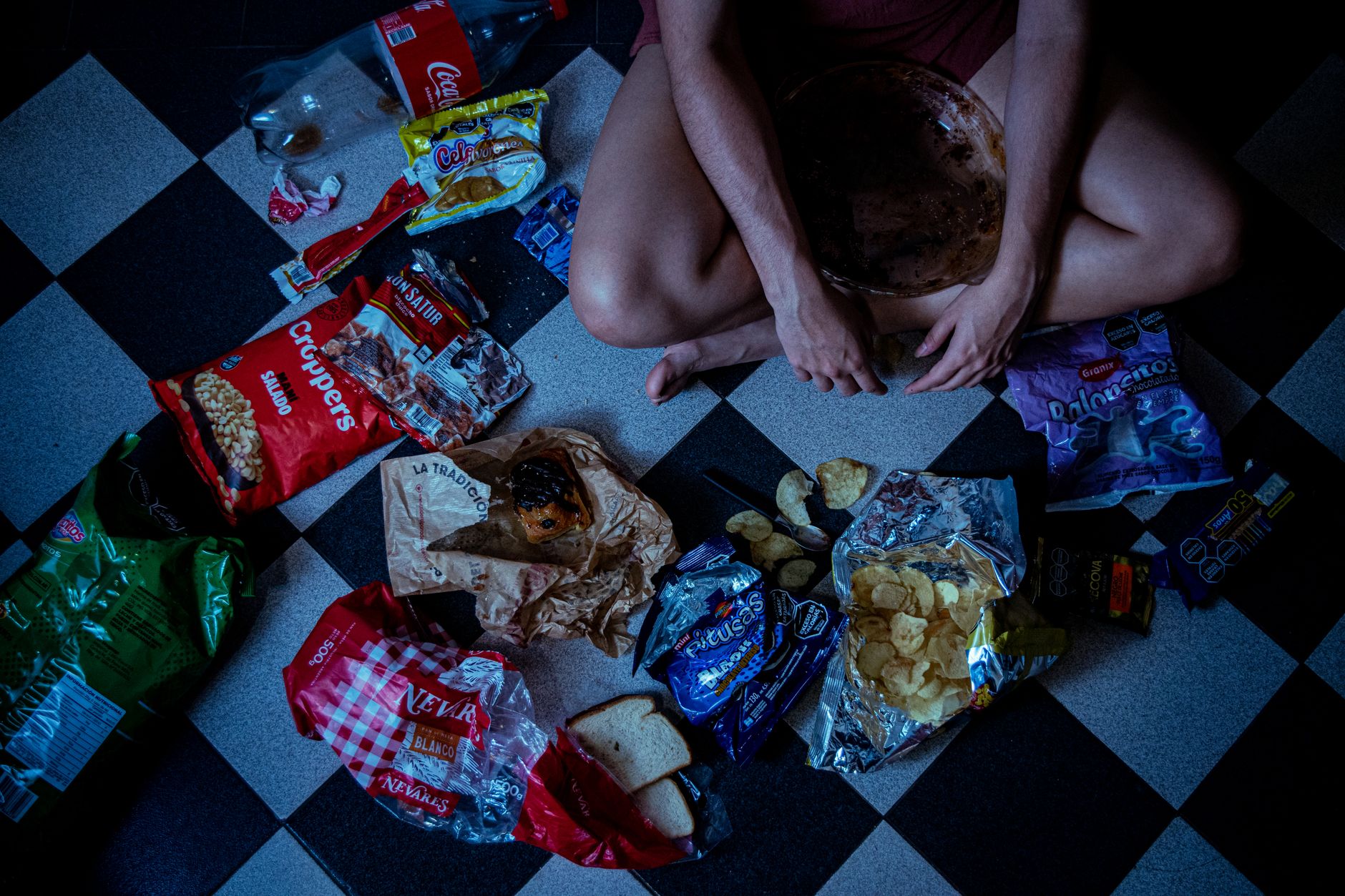 Man sitting surrounded by assorted snacks and packaging on a checkered floor, depicting a binge-eating scene.