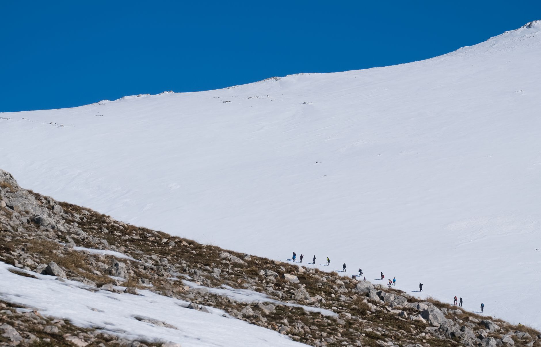 A line of trekkers ascend a snowy mountain under a clear blue sky.