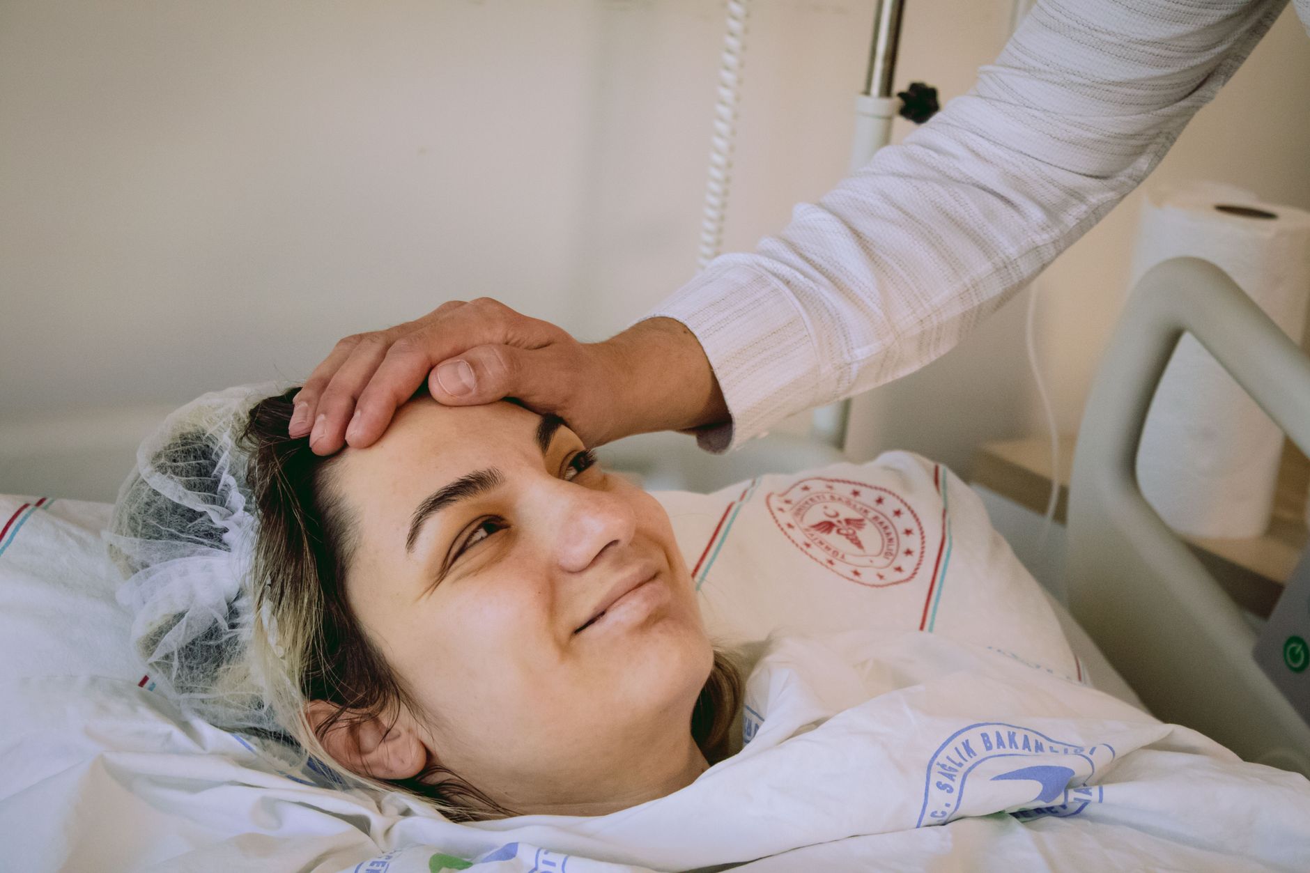 Smiling woman in hospital bed with supportive hand on forehead, conveying warmth and care.