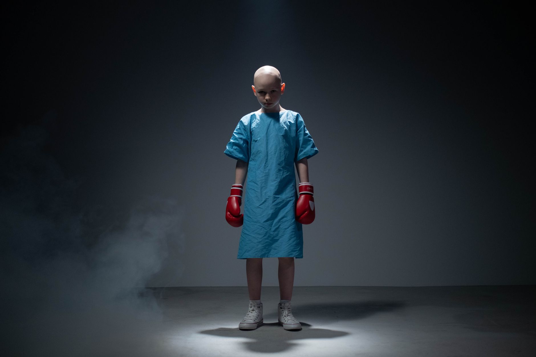 A young child in a hospital gown wears boxing gloves, symbolizing strength and resilience.